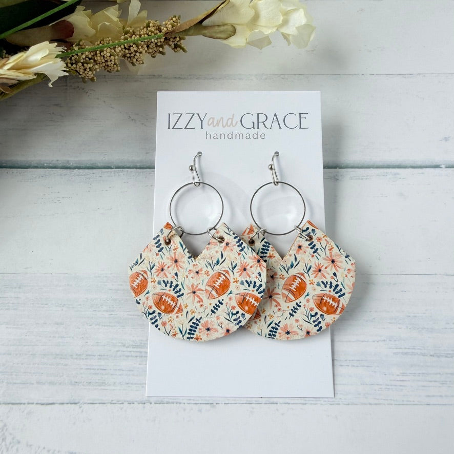 A pair of handmade dangle earrings displayed on a white card labeled 'IZZY and GRACE handmade,' placed on a white wood surface with dried flowers in the corner. The earrings have silver-tone hoop tops and fan-shaped fabric drops in a floral print with brown football motifs.