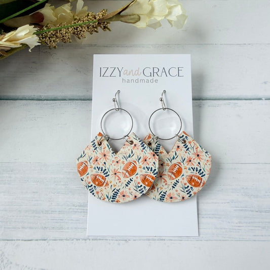 A pair of handmade dangle earrings displayed on a white card labeled 'IZZY and GRACE handmade,' placed on a white wood surface with dried flowers in the corner. The earrings have silver-tone hoop tops and fan-shaped fabric drops in a floral print with brown football motifs.