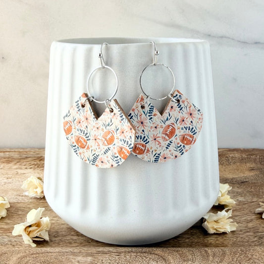 A pair of silver hoop earrings with fan-shaped dangles printed with brown footballs and blue floral motifs on a white background, displayed on a white ribbed ceramic mug.