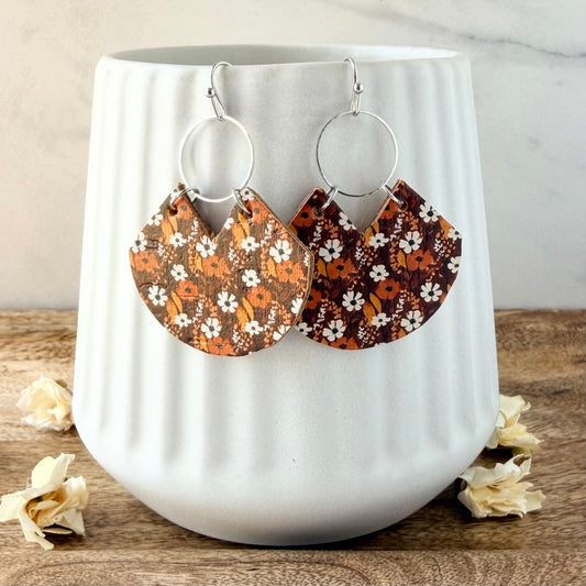 A pair of bohemian-style dangle earrings with silver-tone hoops and fan-shaped charms featuring a brown, orange, and white floral pattern, displayed on a white ceramic vase.
