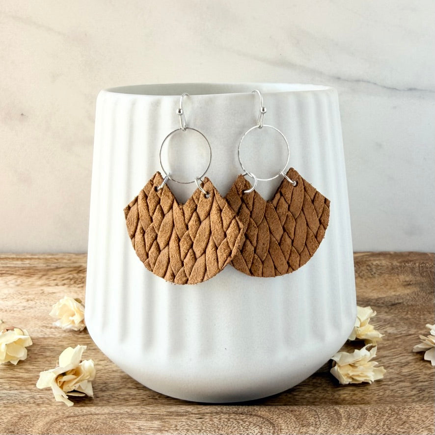 A pair of handmade brown leather dangle earrings with a textured, woven-look pattern and silver-tone hoop backs, displayed on a white ribbed ceramic mug.
