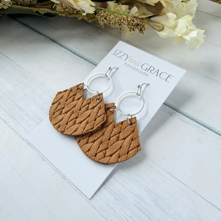 A pair of mustard woven leather dangle earrings with silver hoop backings, displayed on a white card labeled 'IZZYwith GRACE handmade' on a light wood surface with dried flowers in the background.