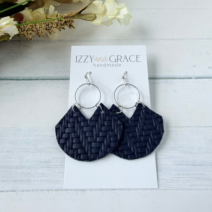 A pair of handmade dangle earrings with dark navy woven leather fan-shaped drops and silver hoop earrings, displayed on a white card labeled 'IZZY and GRACE handmade' on a light wood background with dried flowers.