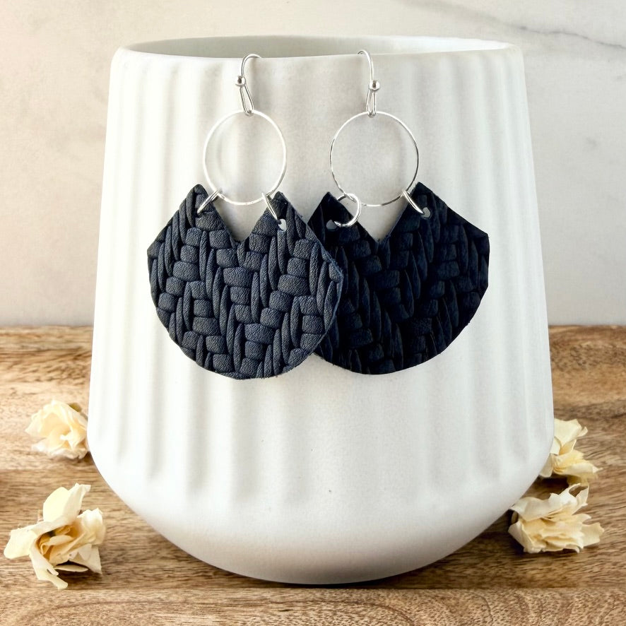 Navy blue woven leather dangle earrings with silver-tone hoop backings, displayed on a white ribbed ceramic vase on a wooden surface with dried flowers.