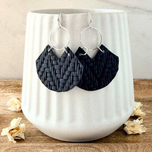 Navy blue woven leather dangle earrings with silver-tone hoop backings, displayed on a white ribbed ceramic vase on a wooden surface with dried flowers.