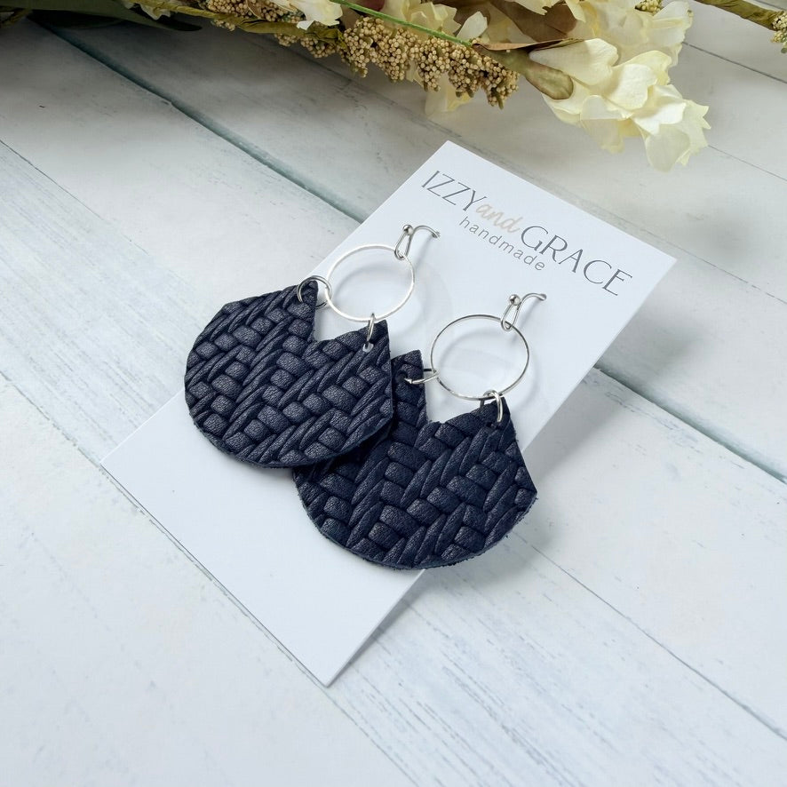 A pair of dark navy blue woven leather fan shaped drop earrings with silver-tone hoop, displayed on a white card labeled 'IZZYwith GRACE handmade' on a white wood background.