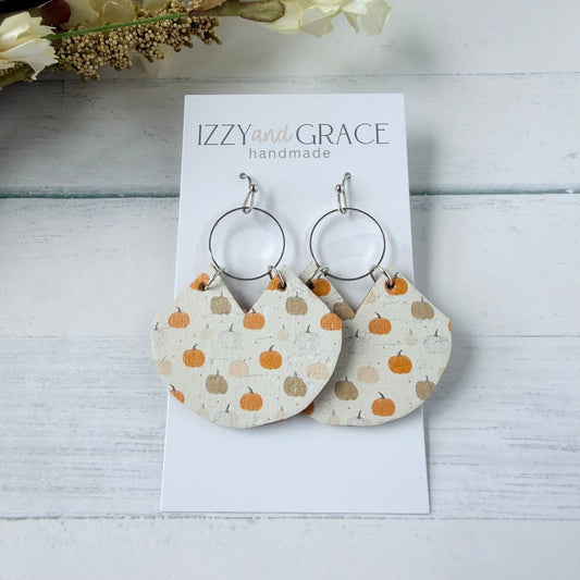 A pair of handmade dangle earrings displayed on a white card labeled 'IZZY and GRACE handmade.' The earrings have silver-tone hoops and semi-circular drops covered in a cream fabric print with orange pumpkins and small brown stems.