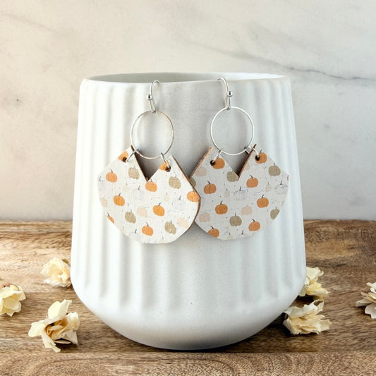 A pair of handmade dangle earrings with silver-tone hoops and fan-shaped charms printed with orange and gray pumpkins on a white background, displayed on a white ribbed ceramic vase.
