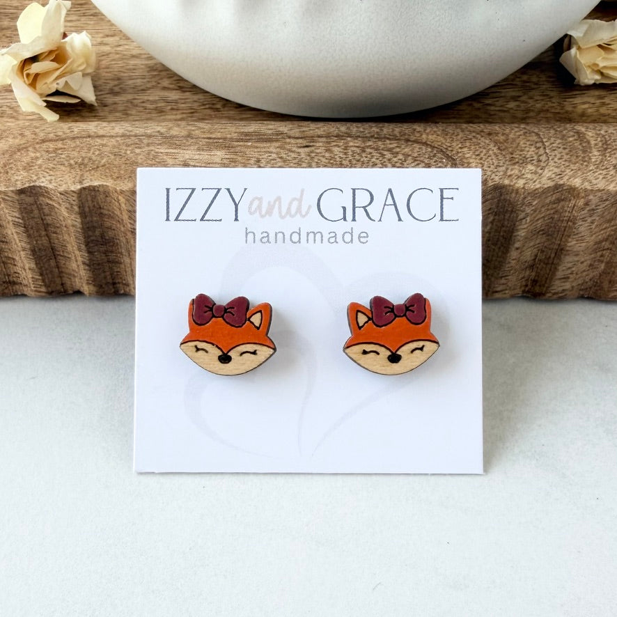 A pair of small fox face stud earrings on a white display card reading 'IZZY and GRACE handmade,' placed on a light surface with a wood block and white bowl in the background.