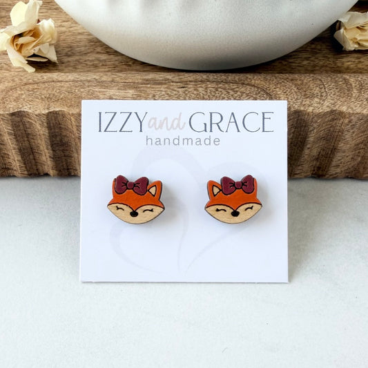 A pair of small fox face stud earrings on a white display card reading 'IZZY and GRACE handmade,' placed on a light surface with a wood block and white bowl in the background.