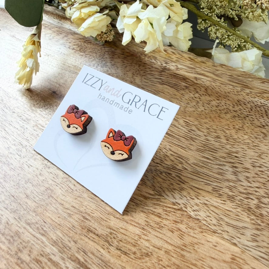 A pair of handmade stud earrings shaped like fox faces with orange fur, brown ears, and red bows, displayed on a white card labeled 'IZZYand GRACE handmade' on a wooden surface with flowers in the background.