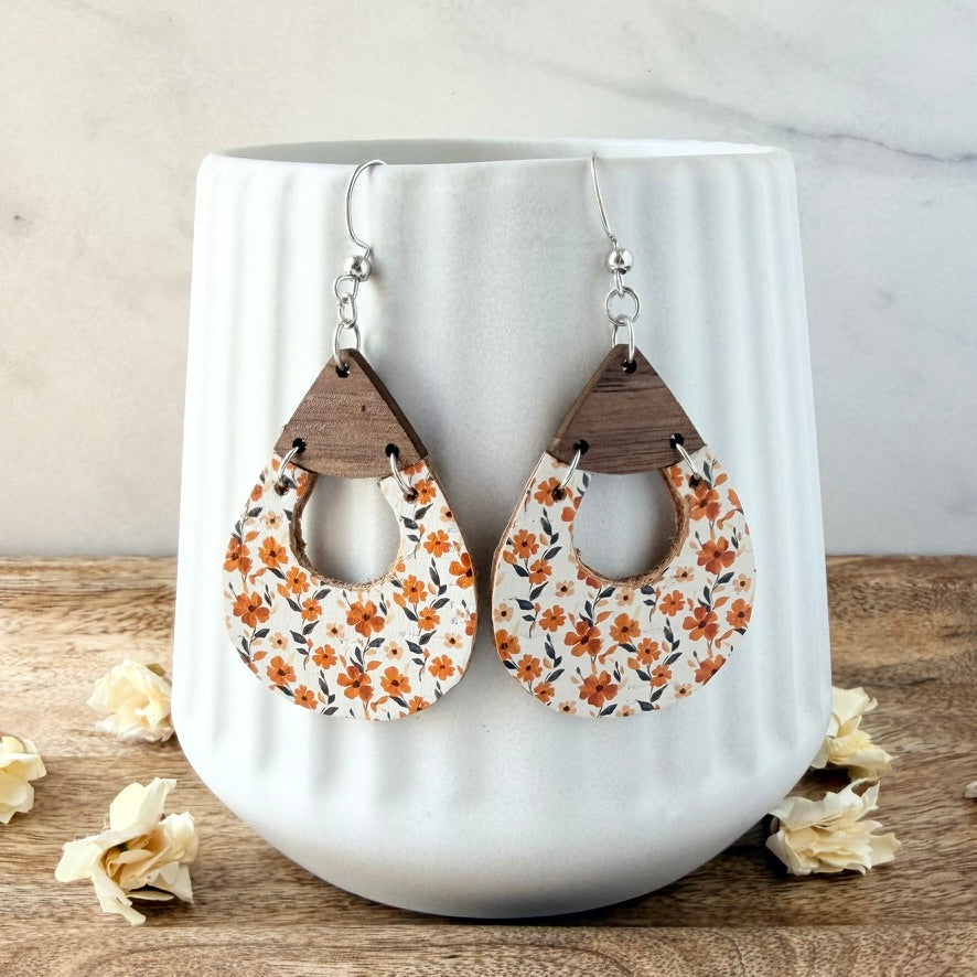 Teardrop-shaped dangle earrings with a white background, orange floral print, and a dark wood top section, hanging in front of a white ribbed ceramic mug on a wooden surface.