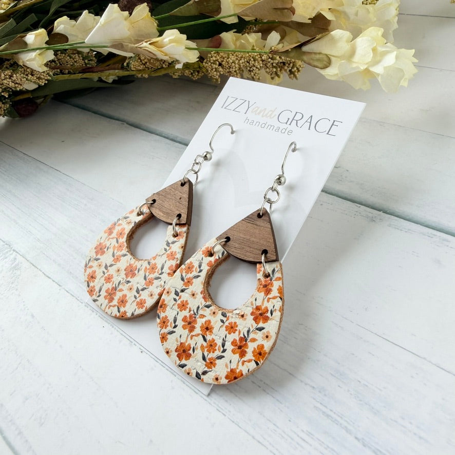 Teardrop-shaped wooden dangle earrings with a white floral print and natural wood accents, displayed on a white card labeled 'Izzy and Grace Handmade' on a white wood surface with dried flowers in the background.