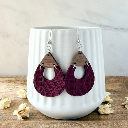 A pair of teardrop-shaped dangle earrings with burgundy embossed leather and triangular wooden tops, displayed on a white ribbed ceramic vase on a wooden surface with dried flowers.
