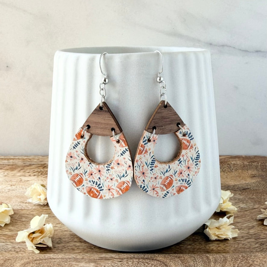 A pair of teardrop-shaped wooden dangle earrings with a floral and football pattern, displayed on a white ribbed ceramic mug on a wooden surface.