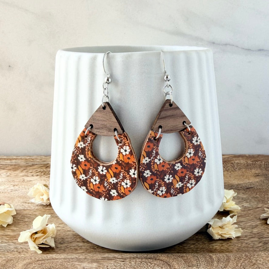 A pair of teardrop-shaped wooden dangle earrings with a brown, orange, and white floral pattern, hanging in front of a white ribbed ceramic vase on a wooden surface.