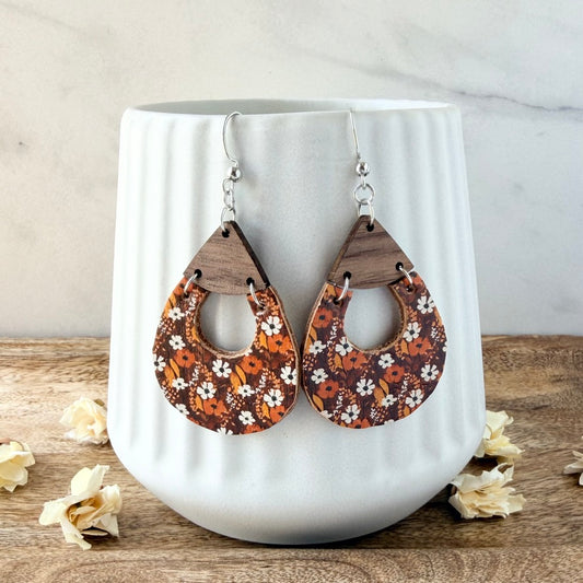 A pair of teardrop-shaped wooden dangle earrings with a brown, orange, and white floral pattern, hanging in front of a white ribbed ceramic vase on a wooden surface.