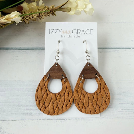 A pair of teardrop-shaped dangle earrings with braided tan leather and dark brown wood accents, displayed on a white card labeled 'IZZYand GRACE handmade' on a light wood background.