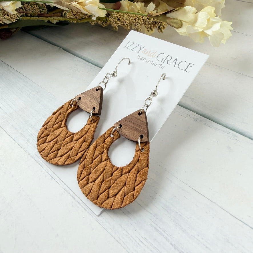 A pair of handmade teardrop-shaped dangle earrings made from textured tan woven leather with a small dark wood accent at the top and silver hooks, displayed on a white card labeled 'Izzy and Grace Handmade' on a white wood background with dried flowers.