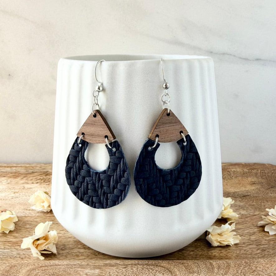 A pair of large teardrop-shaped dangle earrings with dark navy woven leather halves and natural wood top sections, hanging from silver fishhook wires. They are displayed draped over a white ceramic vase on a wooden surface with dried flowers.