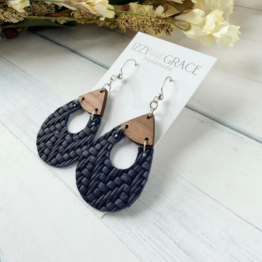 A pair of handmade dangle earrings displayed on a white card. The earrings feature a dark navy blue woven leather lower half, a natural brown wood triangular connector, and silver-tone hooks.