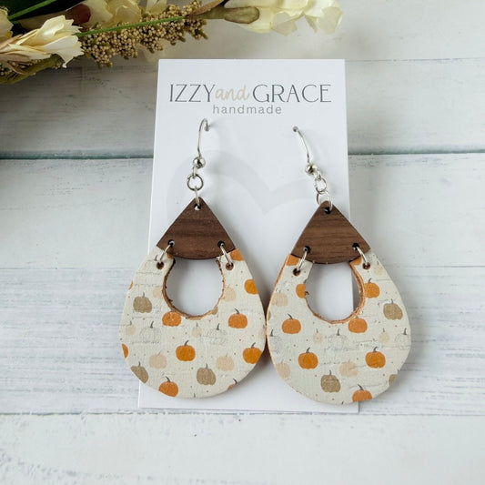 Teardrop-shaped dangle earrings with a pumpkin print on a cream fabric, attached to dark brown wooden triangles and silver hooks, displayed on a white card labeled 'IZZY and GRACE handmade' on a white wood background.