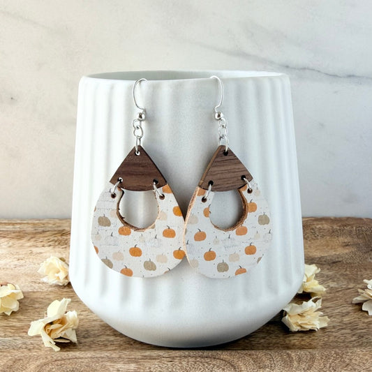 A pair of teardrop-shaped dangle earrings with a printed with small orange and tan pumpkins, attached to dark brown wooden triangles and silver hooks, displayed on a white ceramic vase.