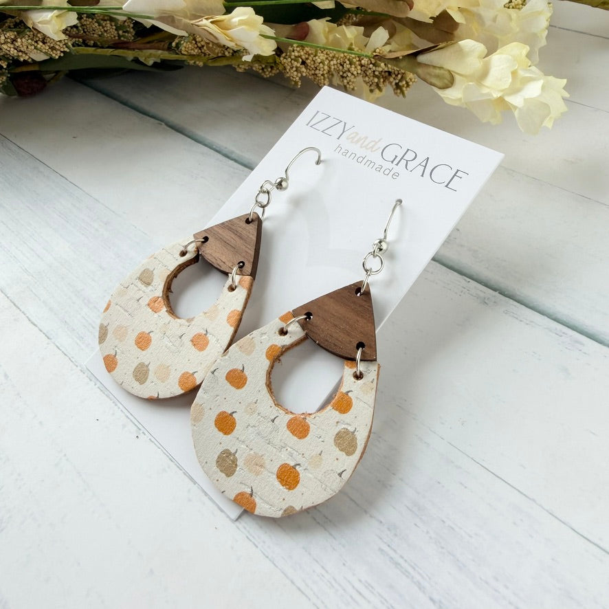 A pair of handmade teardrop-shaped dangle earrings displayed on a white card labeled 'Izzy and Grace handmade.' The earrings have a cream base with orange and brown pumpkin motifs and dark brown wooden top sections, shown on a white wood surface with dried flowers in the background.