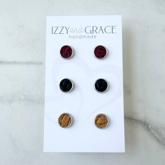 Six round stud earrings displayed on a white card labeled 'IZZY and GRACE handmade,' photographed on a light marble-like surface. The earrings come in burgundy, black, and tan/brown metallic finishes.