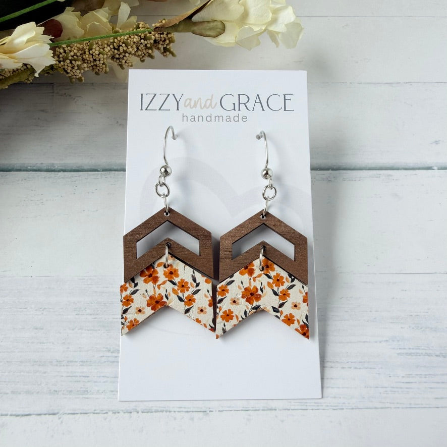 A pair of handmade geometric dangle earrings displayed on a white card labeled 'IZZY and GRACE handmade.' The earrings have brown wooden chevron shapes with a leather bottom featuring orange and yellow floral patterns.