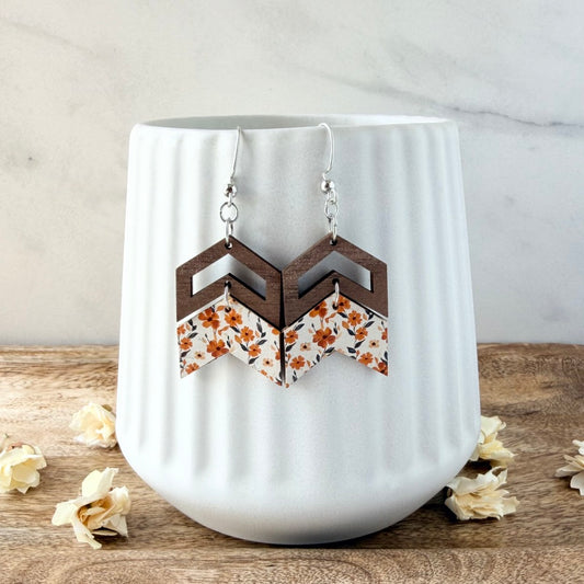 A pair of geometric drop earrings with dark brown wood-look chevron tops and white fabric lower panels printed with orange and yellow floral motifs, hanging in front of a white ribbed ceramic cup on a wooden surface.