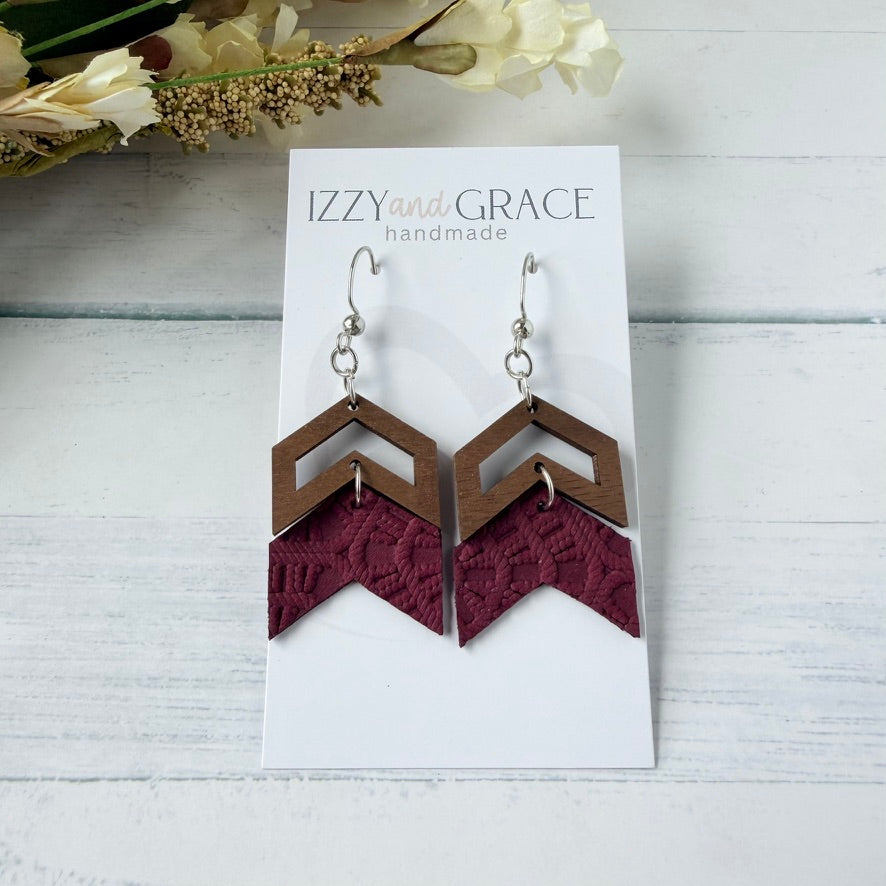 A pair of geometric dangle earrings displayed on a white card labeled 'IZZY and GRACE handmade.' The earrings have silver fishhook wires, brown wooden chevron shapes, and burgundy embossed leather.