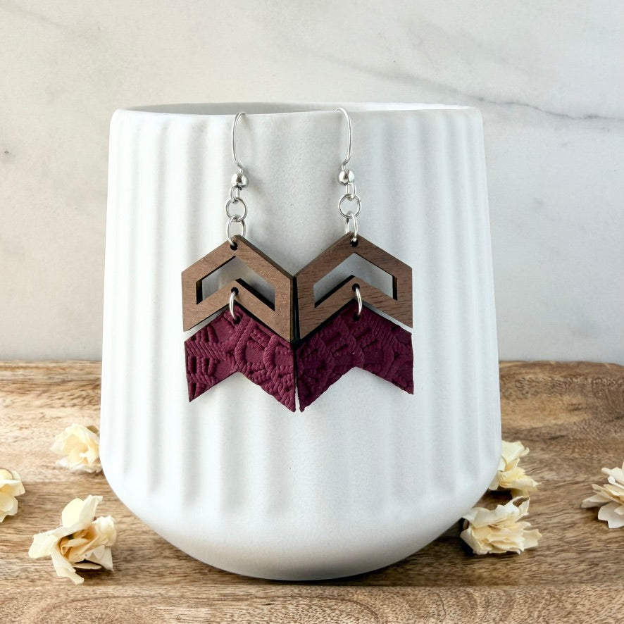 A pair of geometric drop earrings with silver fishhook wires, featuring a medium brown wooden chevron top and a dark burgundy lace chevron bottom, displayed on a white ribbed ceramic cup.