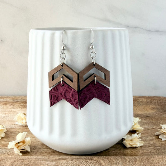 A pair of geometric drop earrings with silver fishhook wires, featuring a medium brown wooden chevron top and a dark burgundy lace chevron bottom, displayed on a white ribbed ceramic cup.