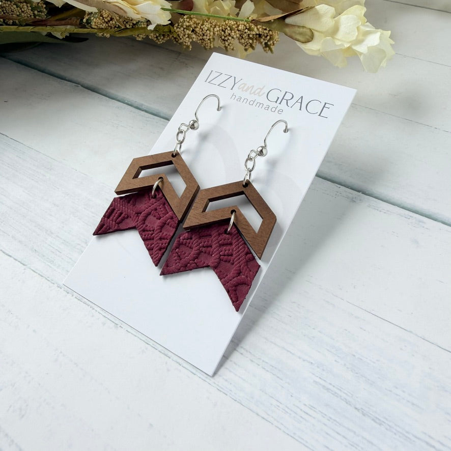 A pair of handmade geometric dangle earrings displayed on a white card. The earrings feature a brown wooden chevron top with a burgundy embossed leather bottom hanging from silver fishhook wires.