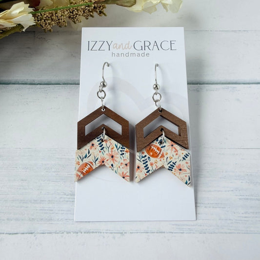 A pair of handmade geometric dangle earrings displayed on a white card labeled 'IZZY and GRACE handmade.' The earrings have silver fishhook wires, dark brown wooden chevron shapes, and lower panels with a floral print in pink, orange, navy, and cream.