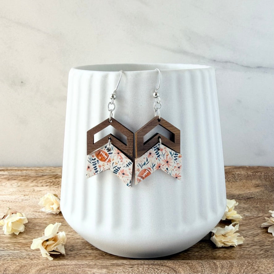 A pair of geometric drop earrings displayed on a white ribbed ceramic vase. The earrings feature dark brown wooden chevron tops and floral patterned dangles in pink, orange, and blue on a white background.