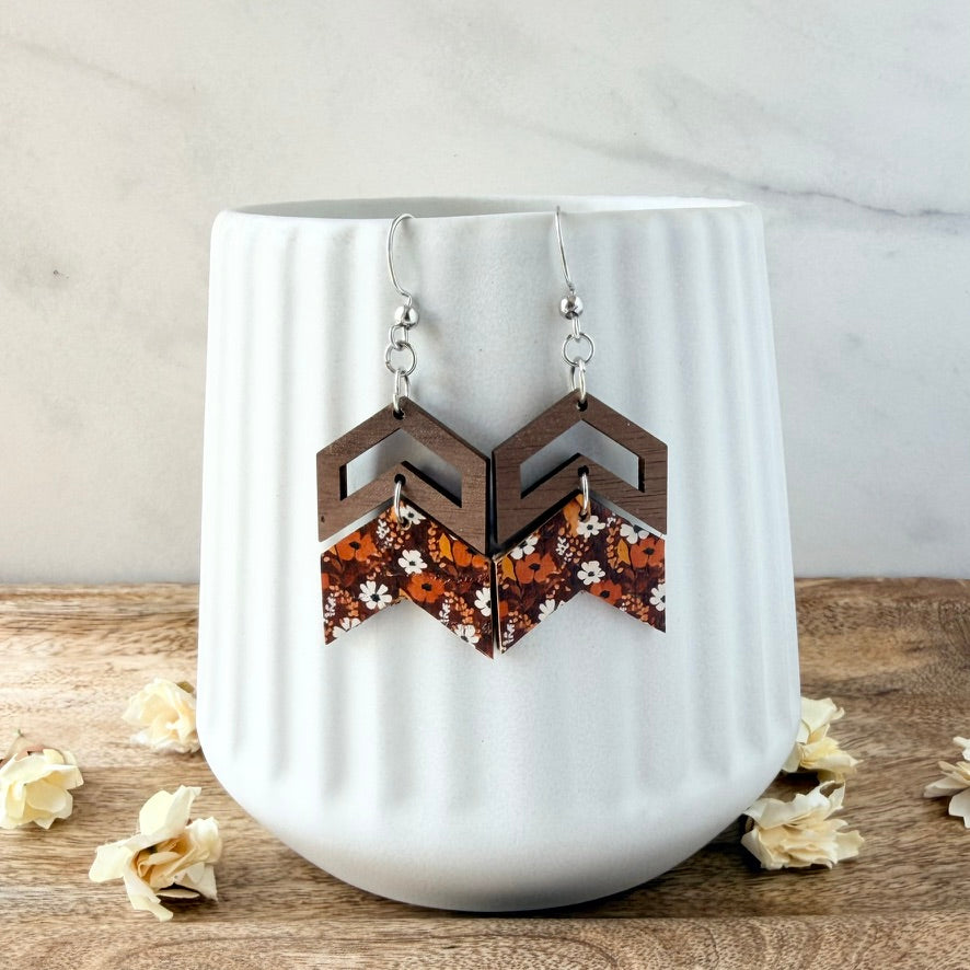 A pair of geometric drop earrings displayed on a white ribbed ceramic vase. The earrings feature dark brown wooden chevron shapes on top and patterned wooden triangles on the bottom with orange, brown, and white floral details.
