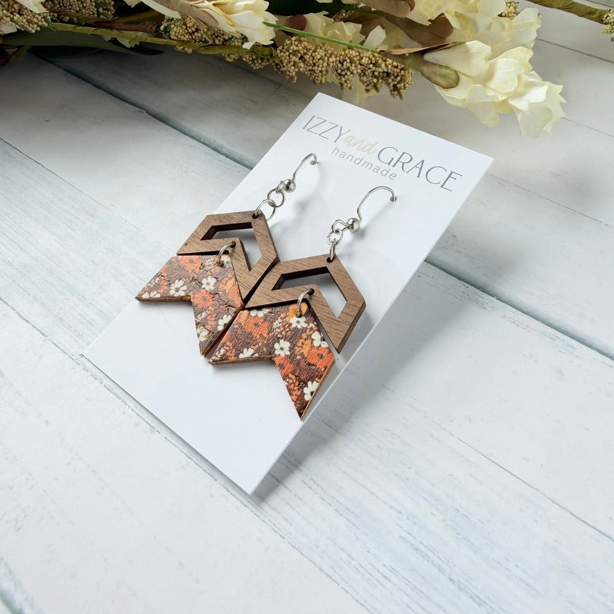 A pair of geometric drop earrings displayed on a white card labeled 'Izzy and Grace handmade.' The earrings have layered brown wood triangles over orange, brown, and white floral pieces with silver fishhook wires.