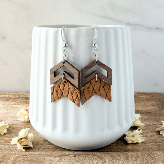 A pair of geometric wooden drop earrings with silver fishhook wires, displayed on a white ribbed ceramic vase. The earrings feature a dark brown wood top and a mustard textured leather bottom.