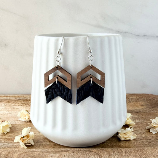 A pair of geometric dangle earrings displayed on a white ribbed ceramic vase. The earrings feature a wooden chevron top, an embossed leather chevron bottom, and silver-toned fishhook wires.