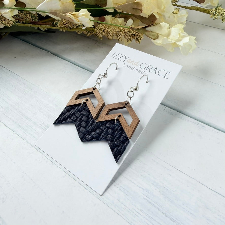 A pair of geometric dangle earrings displayed on a white branded card. The earrings have silver-tone hooks, light natural wood chevron shapes, and dark navy woven leather chevron bottoms.