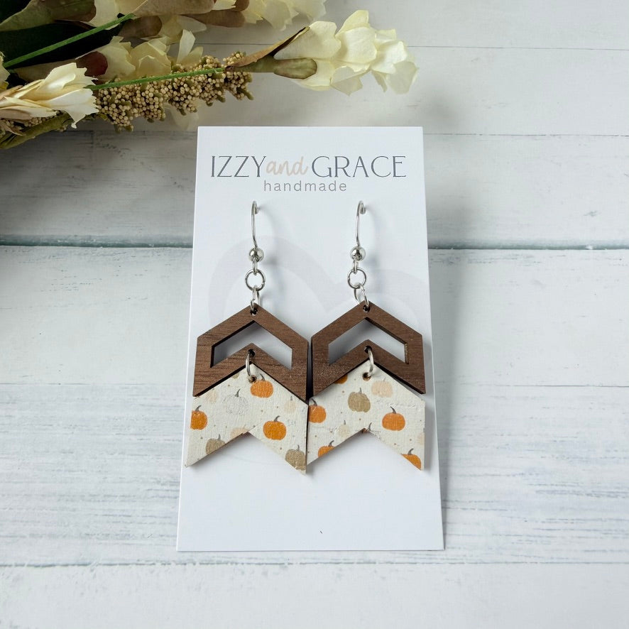 A pair of handmade geometric dangle earrings displayed on a white card labeled 'IZZYand GRACE handmade.' The earrings have silver fishhook wires and layered chevron shapes: dark brown wood on top and a cream fabric lower section with small orange and tan pumpkins.