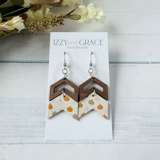 A pair of handmade geometric dangle earrings displayed on a white card labeled 'IZZYand GRACE handmade.' The earrings have silver fishhook wires and layered chevron shapes: dark brown wood on top and a cream fabric lower section with small orange and tan pumpkins.
