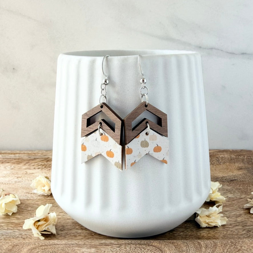 A pair of geometric dangle earrings displayed on a white ribbed ceramic vase. The earrings feature a dark brown wooden geometric top and a white chevron bottom printed with orange pumpkins and small green leaves.