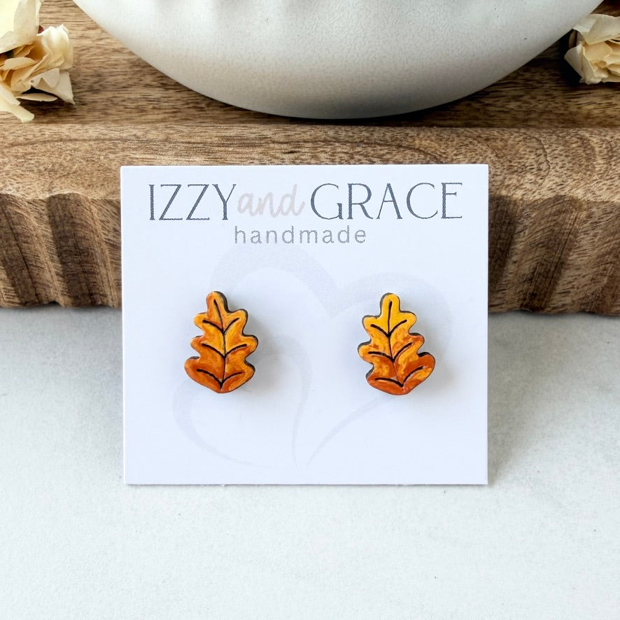 A pair of orange and yellow leaf-shaped stud earrings displayed on a white card that reads 'IZZY and GRACE handmade,' set on a light surface with a wooden board and dried flowers in the background.