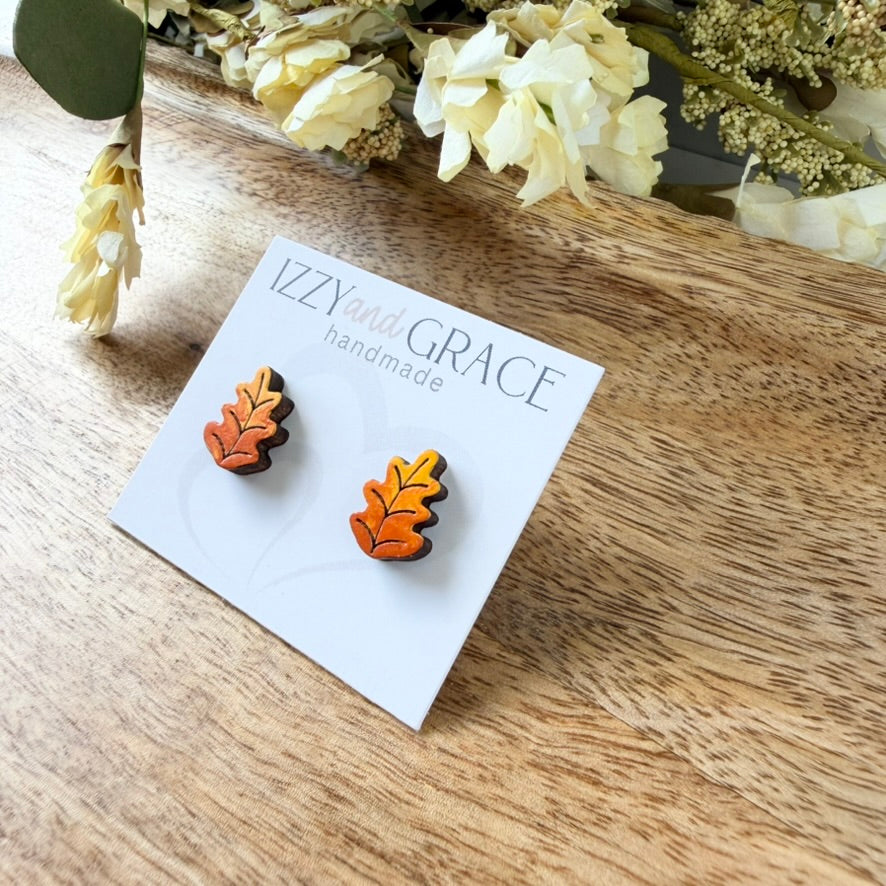 A pair of handcrafted autumn leaf stud earrings in orange, yellow, and brown, displayed on a white card labeled 'Izzie and Grace handmade' on a wooden tabletop with dried flowers in the background.