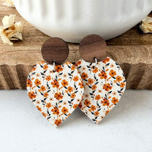 petal-shaped wooden drop earrings with a white leather printed with an orange and floral pattern, featuring round dark brown wooden studs, displayed on a rustic wooden board with a white ceramic bowl in the background.