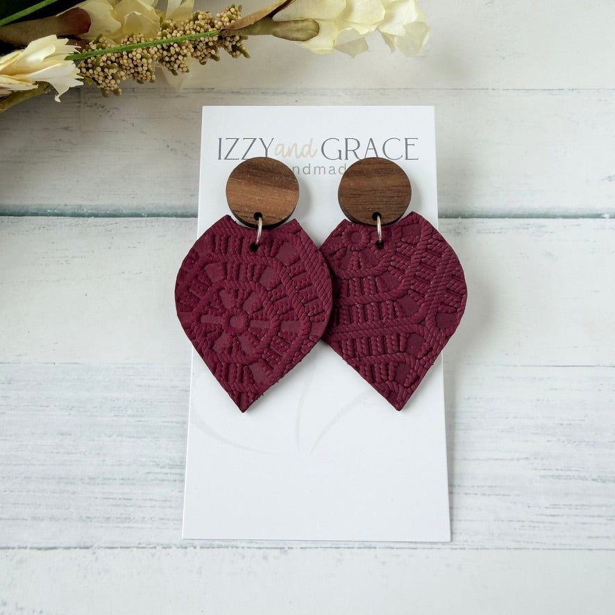 A pair of handmade drop earrings with textured burgundy leather petal-shaped bottoms attached to round wooden studs, displayed on a white card labeled 'IZZY and GRACE handmade' on a white wood background.
