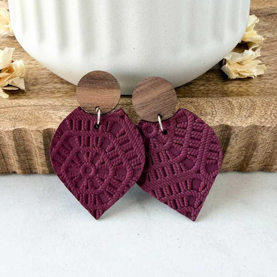 A pair of bohemian-style dangle earrings with textured, embossed burgundy petal-shaped leather bottoms and circular wooden studs, displayed on a wooden board with a white ceramic mug in the background.